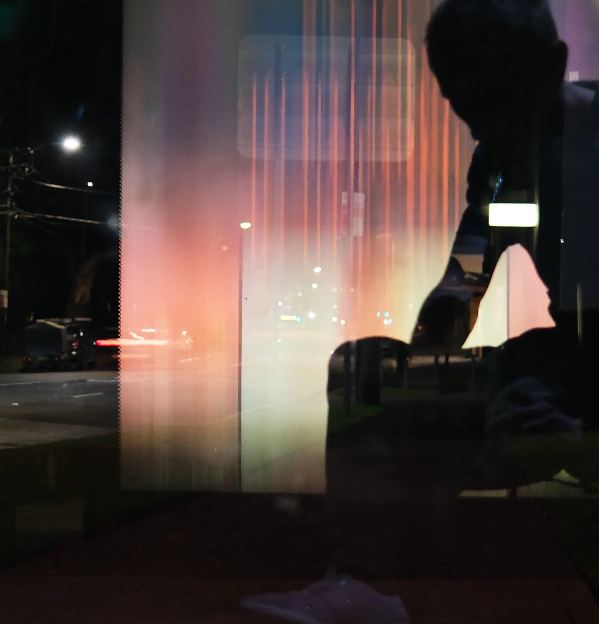 A person's silhouette is reflected in a window at night. Street lights and vehicles illuminate the scene outside, creating colourful vertical streaks on the glass.