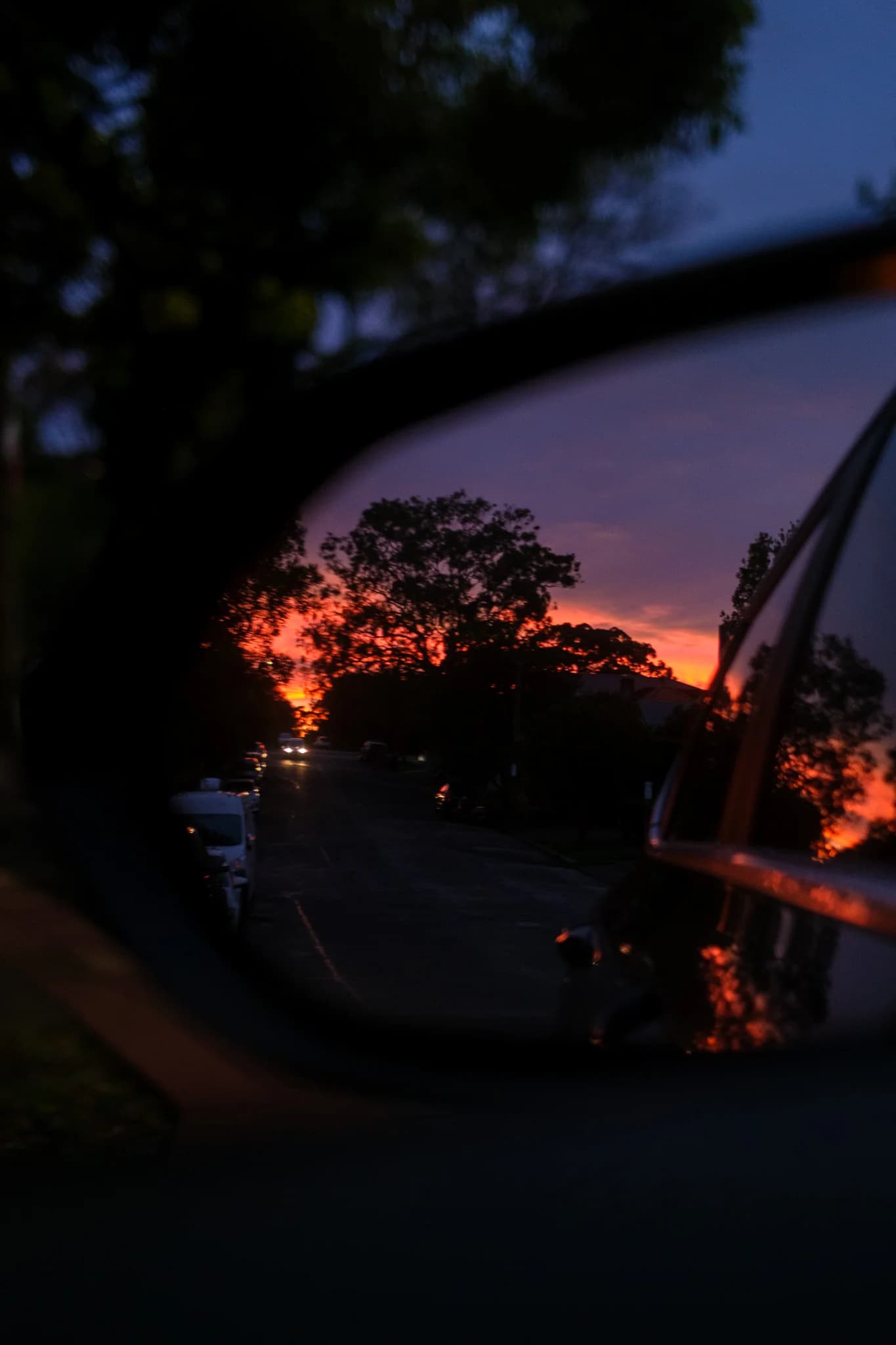 A vibrant orange and purple sunset reflects in a car's side mirror. Silhouetted trees and streetlights are visible down the road.