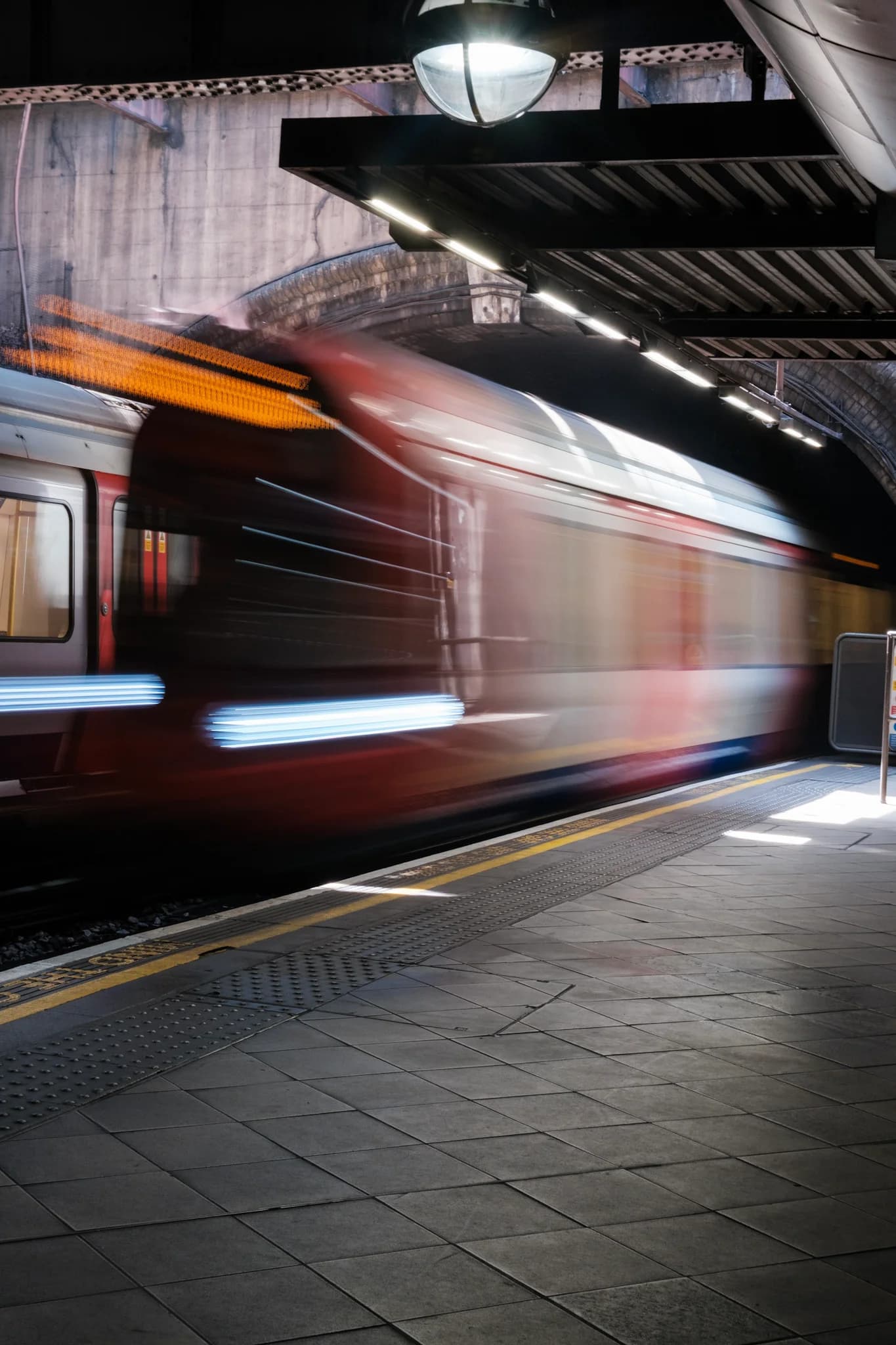 A subway train blurs past an underground platform, creating a sense of motion. Overhead lights illuminate the concrete tunnel and tiled ground.