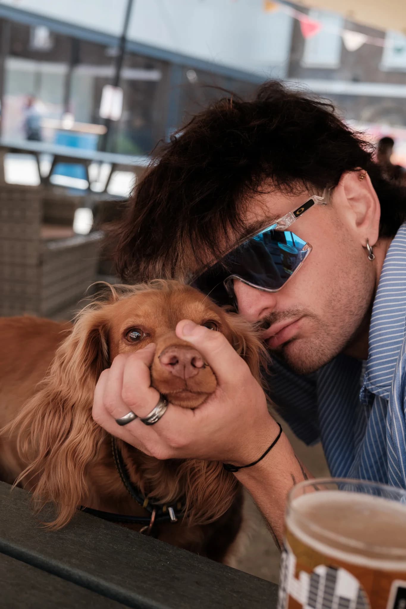 A man wearing reflective sunglasses gently cups the face of a brown spaniel-type dog. A pint glass sits on the wooden table in the foreground.