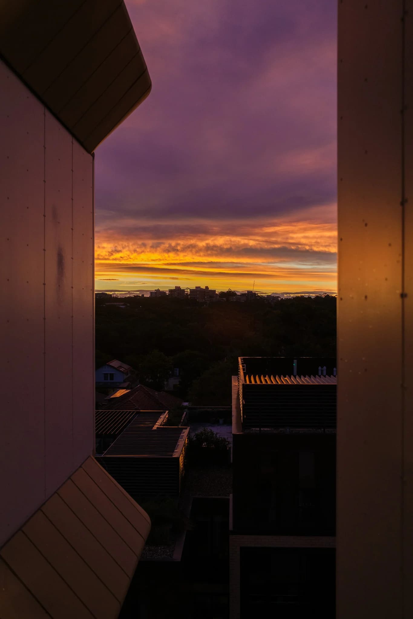 A vibrant purple and orange sunset over a distant skyline, framed by the modern architecture of nearby buildings.