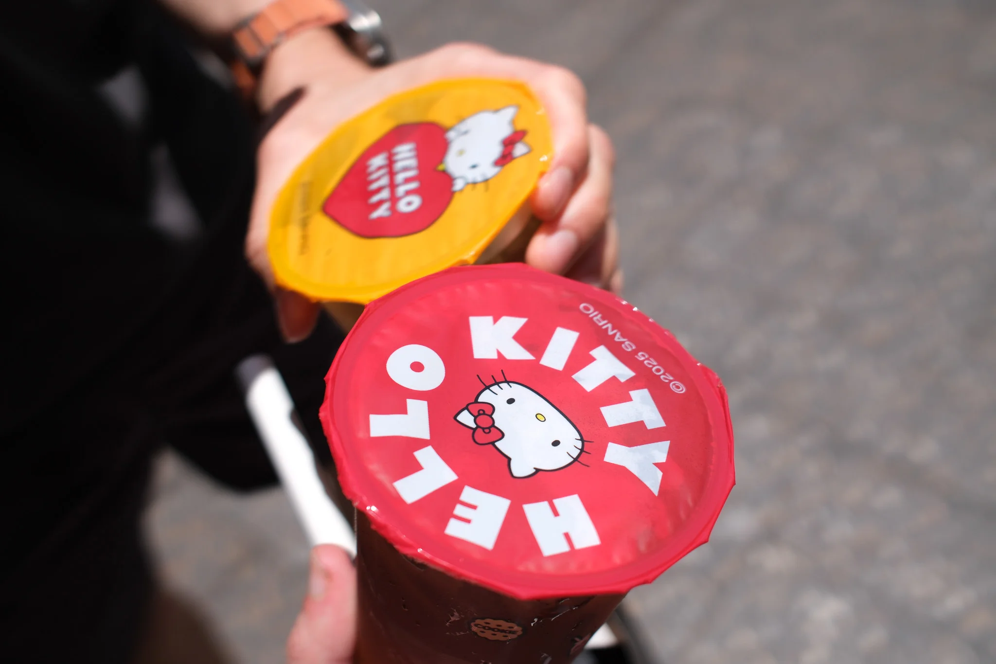 A person holds two bubble tea cups sealed with brightly coloured Hello Kitty branded film. The drinks are displayed outdoors against a blurred paved background.