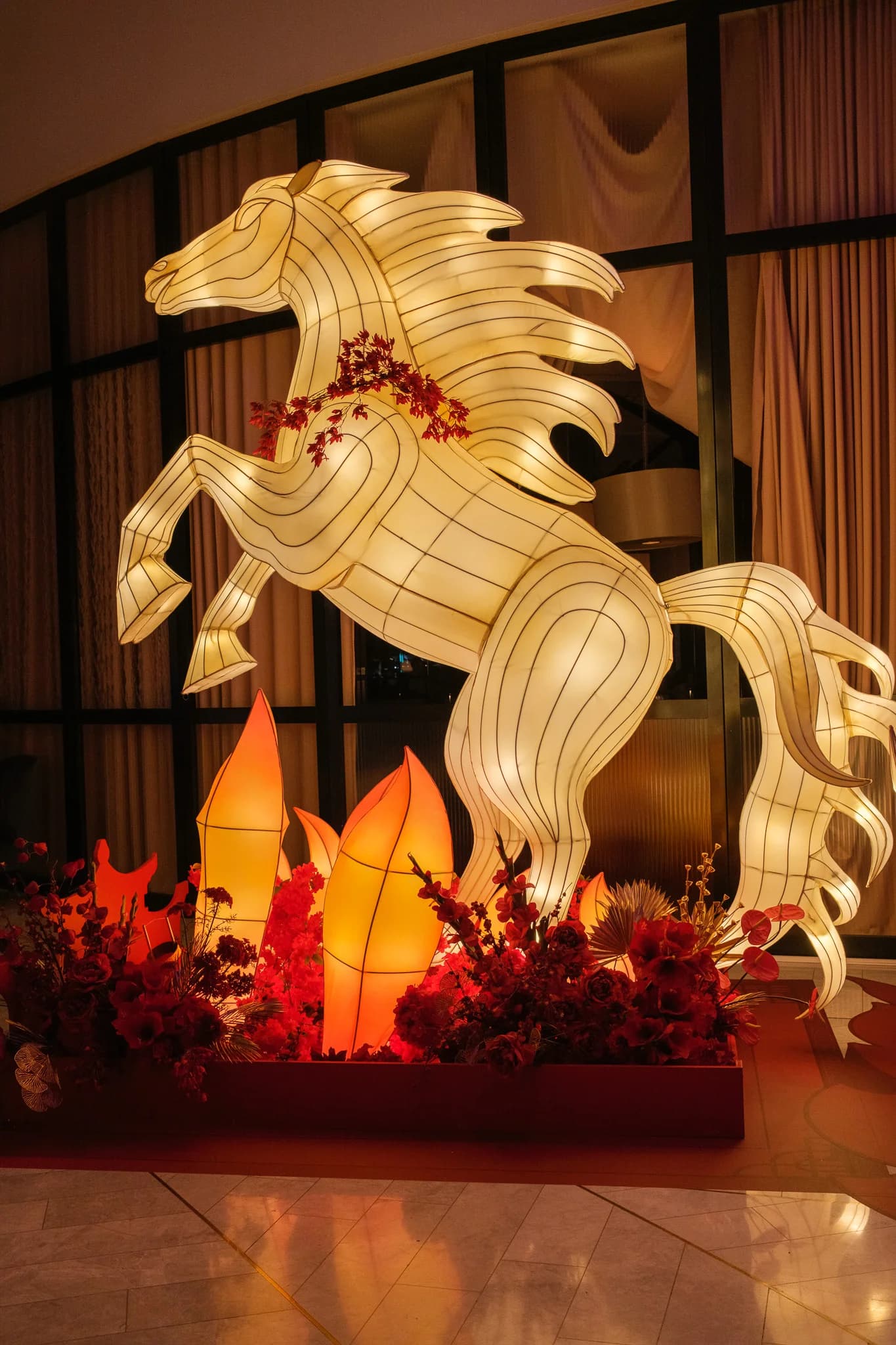 A glowing, rearing horse lantern is displayed indoors with vibrant red flowers and flame-like lights at its base. The intricate wire and fabric construction creates a striking festive centerpiece.