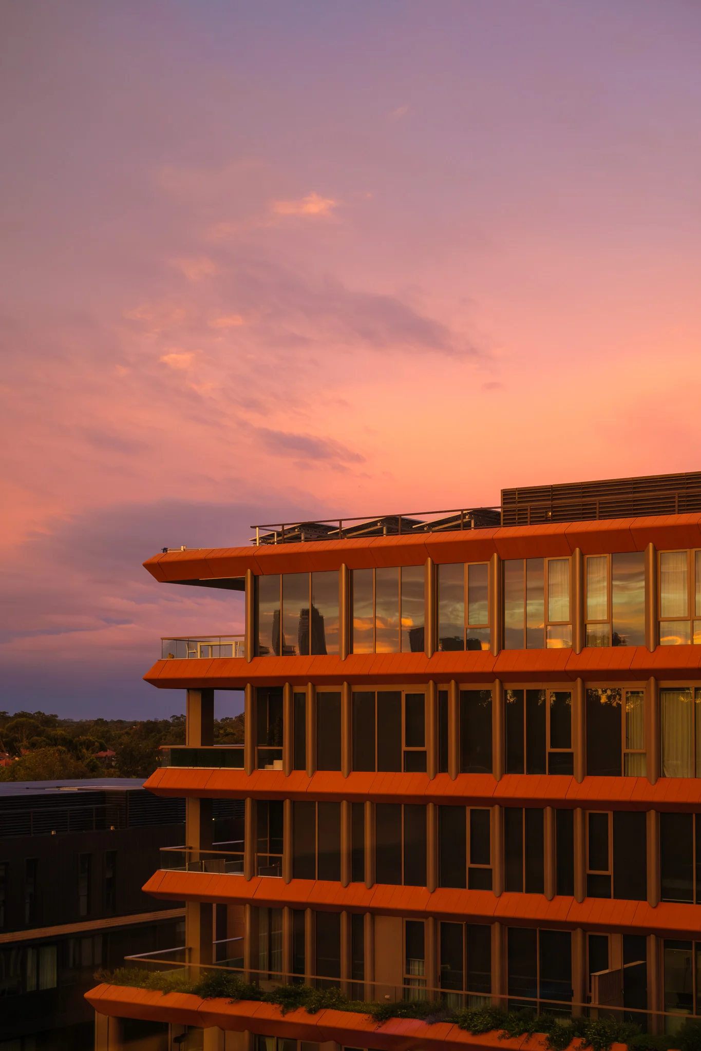 A striking modern building with bright orange accents is illuminated by a vibrant pink and purple sunset. The large glass windows reflect the surrounding cityscape and colourful sky.